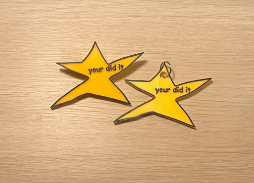 May include: Two yellow star-shaped charms with black outlines and the text "your did it". One charm has a silver keyring attached. The charms are on a light brown wooden surface.