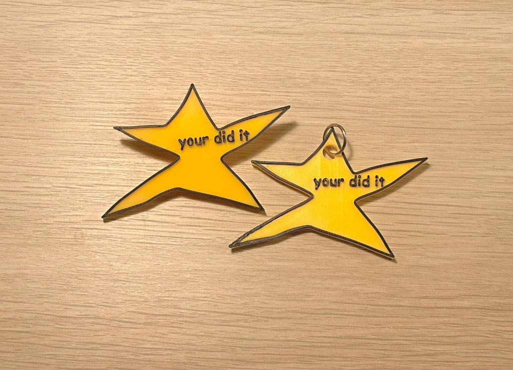 May include: Two yellow star-shaped charms with black outlines and the text "your did it". One charm has a silver keyring attached. The charms are on a light brown wooden surface.
